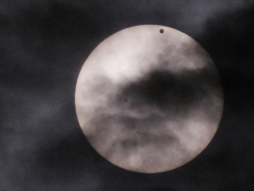 The Sun is silver almost like the moon while a black dot (Venus) and clouds pass in front of it.